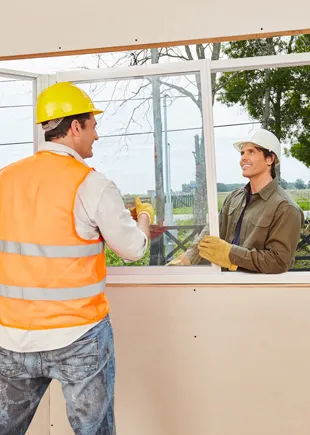 Two fitters in hard hats easing a window frame into a stud opening from either side