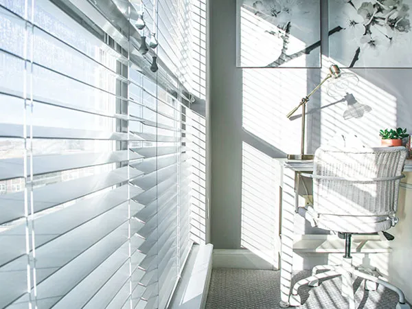 Venetian blinds angled across a home office window, desk and chair beside them in daylight