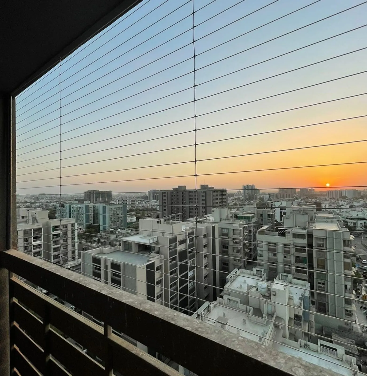Balcony rail with horizontal steel cables strung across it, city rooftops at sunset behind