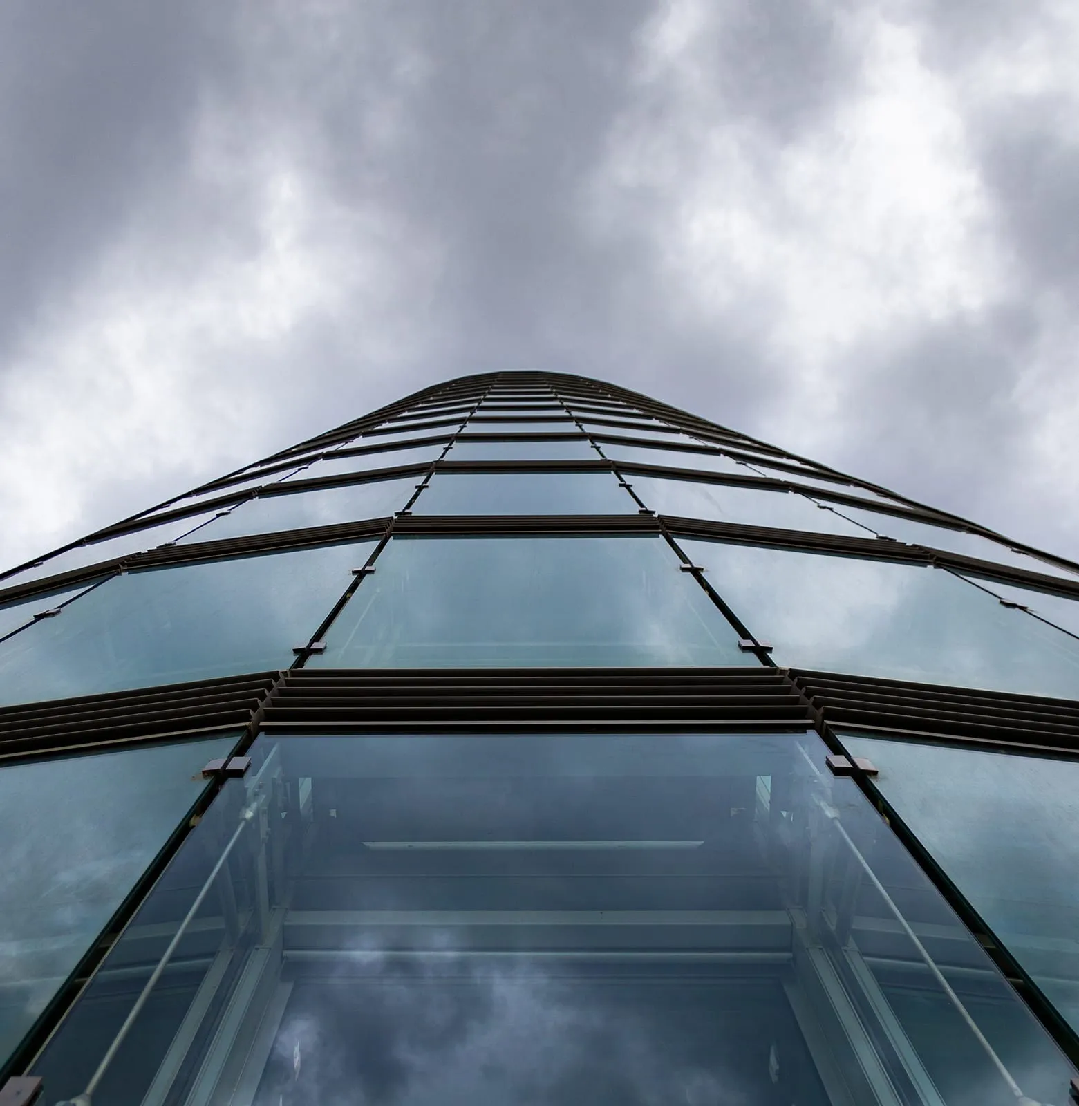 Curved glass office tower seen from its base, panels sweeping upward against grey cloud