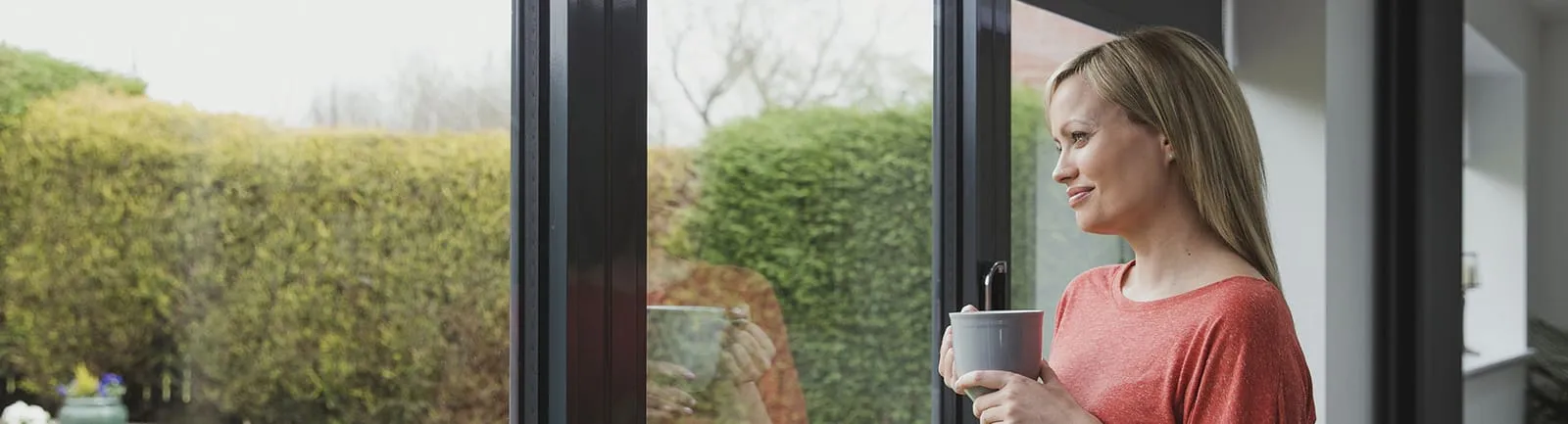 Woman holding a cup beside tall glazed doors, looking out at a garden on an overcast day