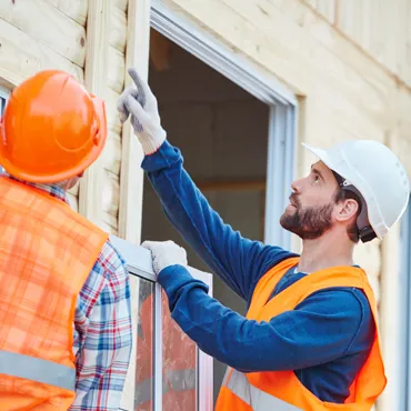Two fitters in hi-vis vests and hard hats lifting a window unit into a prepared opening