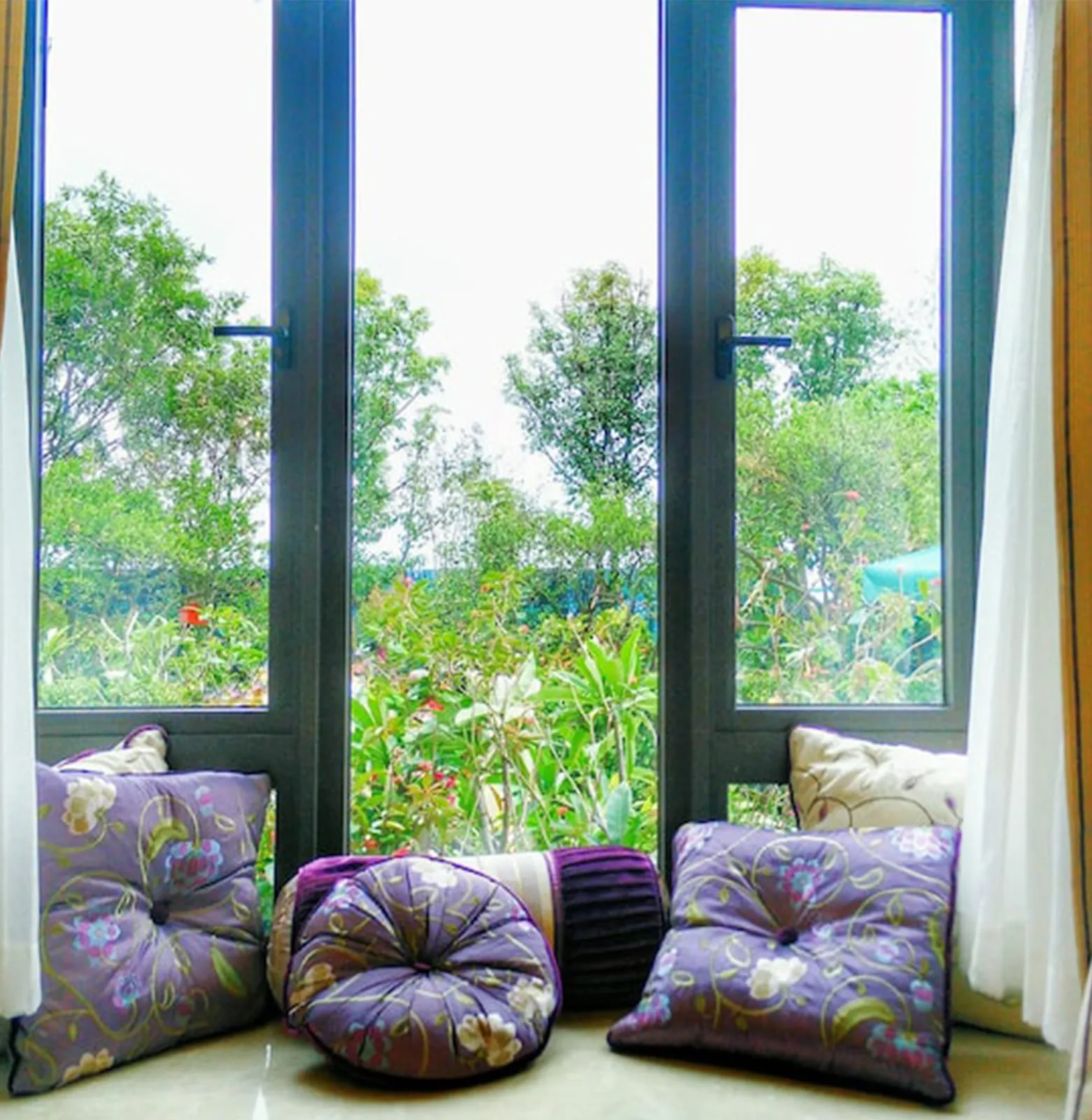 Pair of white casement windows standing open above a cushioned window seat, greenery outside