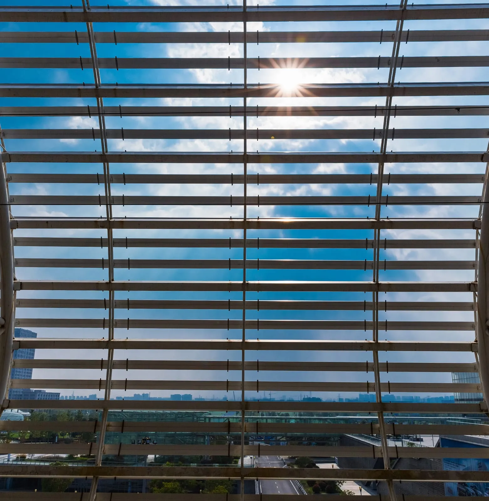Grid of louvred sunshade blades photographed against the sun, sky visible between them